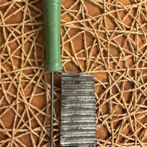 Può includere: Un utensile da cucina vintage con un manico in legno verde e una lama in metallo ondulato. Il manico è cilindrico e verniciato di verde tenue. La lama è rettangolare con creste parallele. L'utensile è progettato per tagliare o grattugiare.