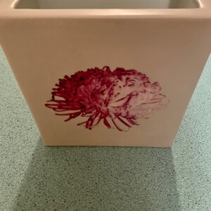 Puede incluir: Un jarrón de cerámica blanco con un diseño de flores rosa y blancas. El jarrón es cuadrado y tiene una abertura ancha.