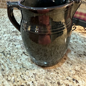May include: A dark brown ceramic pitcher with a wide mouth and a handle. The pitcher has a glazed finish and a slightly textured surface.