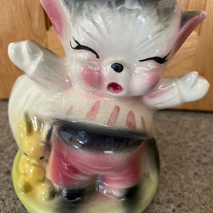 Puede incluir: Una figurita de cerámica de un gato llorando que lleva pantalones rojos y una camisa a rayas blancas y negras. El gato sostiene un pequeño conejito amarillo en su pata izquierda. La figurita está de pie sobre una base verde.