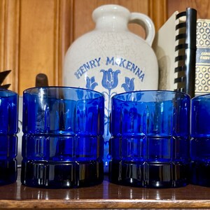 May include: Four cobalt blue glass tumblers with a square patterned design. The tumblers are arranged in a row on a wooden surface.