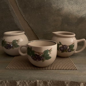 Puede incluir: Tres tazas de cerámica blanca con borde negro y diseño de grosellas negras. Las tazas están sobre un mantel individual tejido marrón.