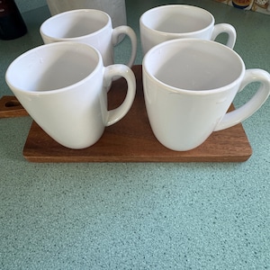 Puede incluir: Cuatro tazas de café de cerámica blanca con asas curvas dispuestas sobre una pequeña tabla de madera rectangular. Las tazas tienen un diseño sencillo y moderno, sobre una encimera azul claro moteada.