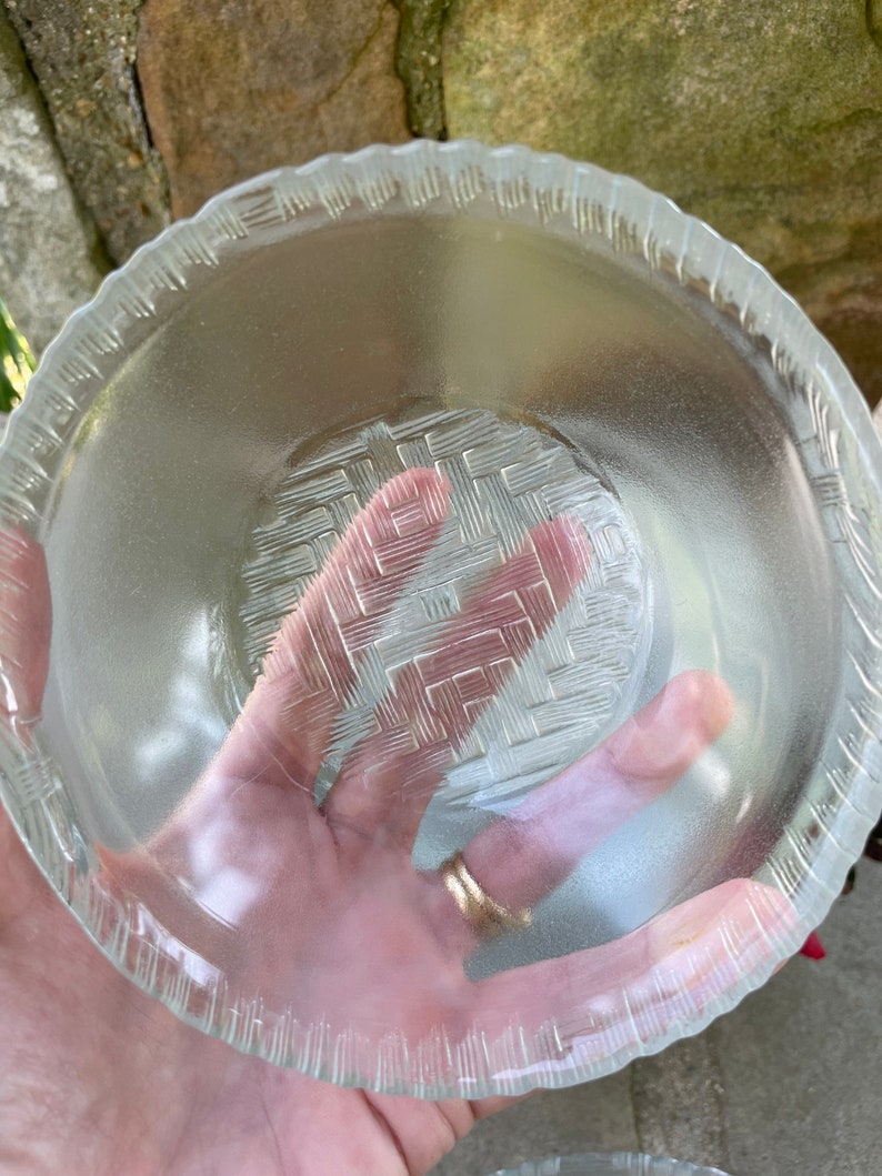 May include: Clear glass bowl with a textured, woven pattern in the center and a scalloped edge. The bowl is transparent, allowing light to pass through. The bowl is likely used for serving food or as a decorative piece.