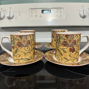 Puede incluir: Cuatro tazas de café de cerámica con un patrón floral en tonos marrones, amarillos y verdes. Las tazas están sobre sus platillos en una encimera negra.