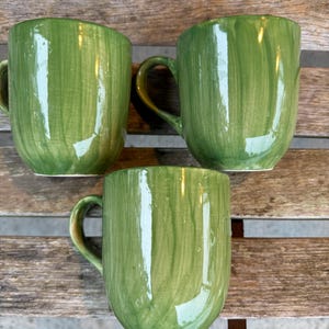 May include: Three green ceramic mugs with a wood grain pattern. The mugs have a rounded shape and a handle on the side.