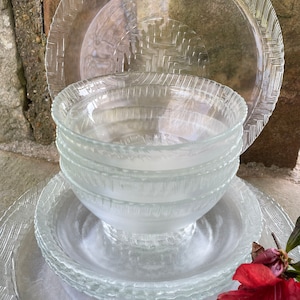 May include: A stack of clear glass dinnerware, including plates and bowls. The plates have a textured, woven-like pattern around the edges. The bowls are stacked in the center. A red flower is in the foreground.