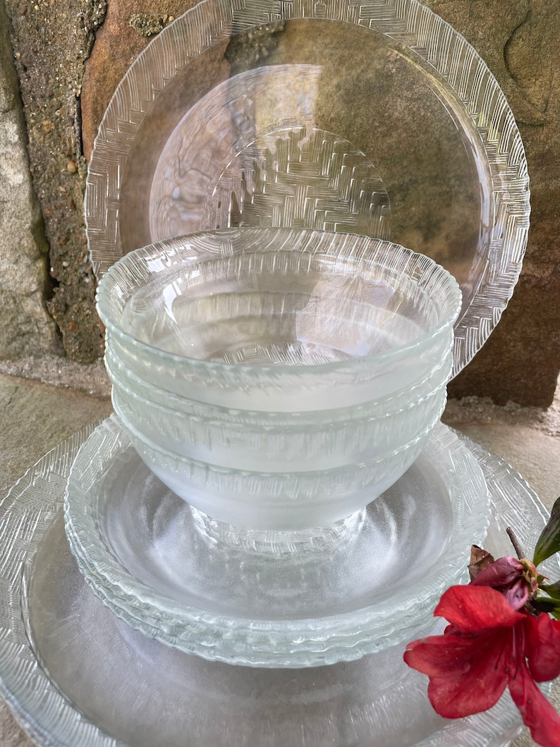 May include: A stack of clear glass dinnerware, including plates and bowls. The plates have a textured, woven-like pattern around the edges. The bowls are stacked in the center. A red flower is in the foreground.