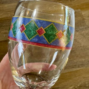 May include: A clear wine glass with a decorative band featuring green, red, and yellow diamond shapes on a blue background. The glass has a rounded bowl and a stem.