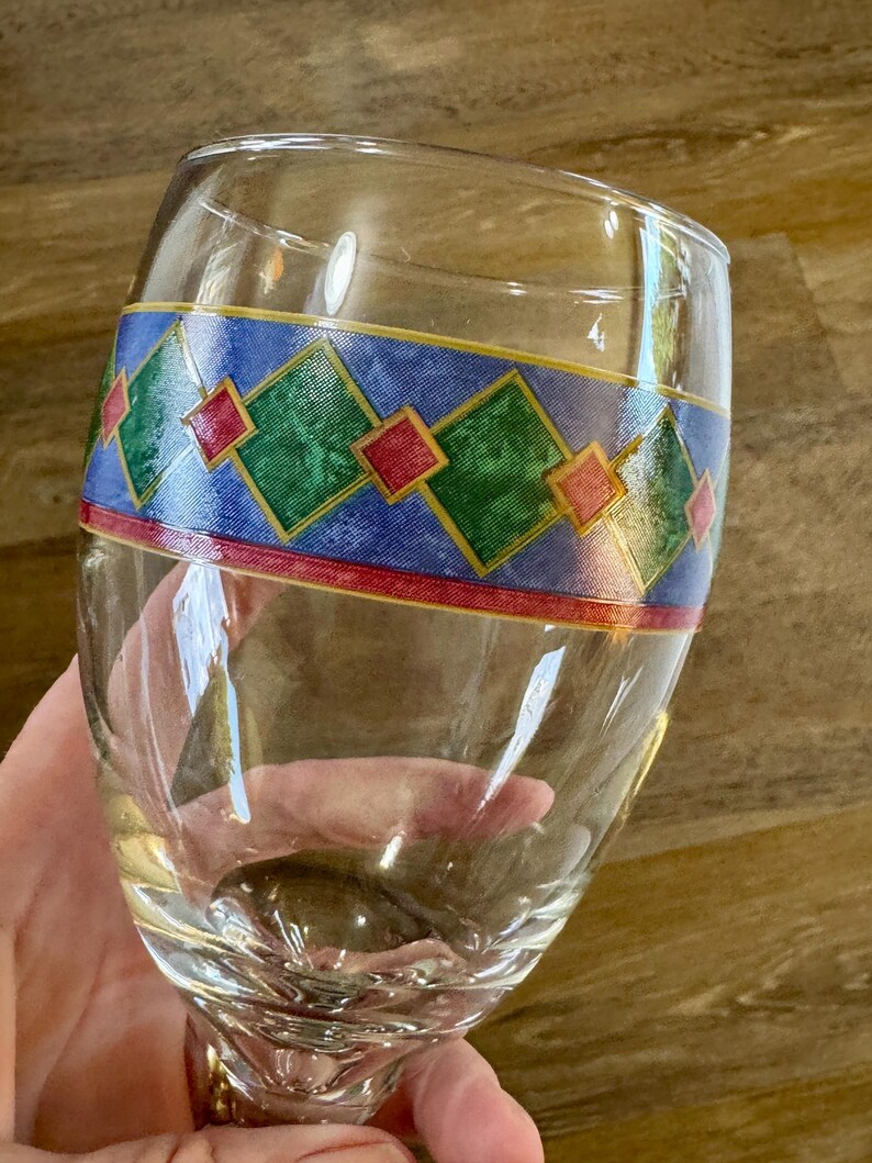 May include: A clear wine glass with a decorative band featuring green, red, and yellow diamond shapes on a blue background. The glass has a rounded bowl and a stem.