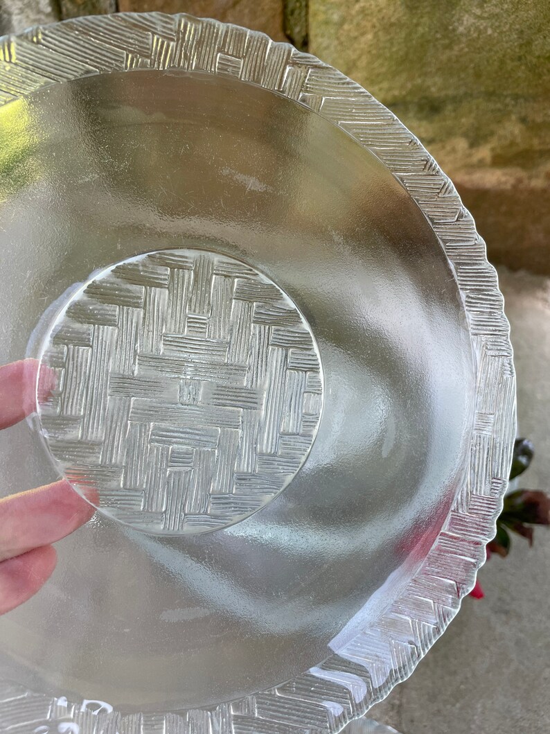 May include: Clear glass serving dish with a woven basket pattern on the base and rim. The dish has a scalloped edge and a circular center. The glass is transparent, allowing light to pass through.