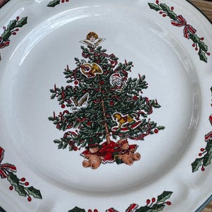 Puede incluir: Plato de Navidad de cerámica blanca con un borde de acebo verde y rojo. En el centro, un árbol de Navidad verde con adornos, un Papá Noel, un ángel y dos osos de peluche.