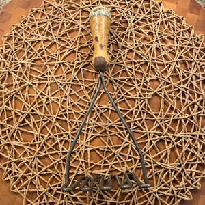 May include: A vintage potato masher with a wooden handle and metal tines. The handle is light brown with a dark brown base and a decorative top. The masher is resting on a woven, circular placemat made of natural fibers.