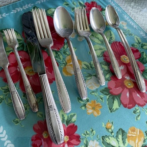 May include: A set of silver-plated cutlery, including a knife, fork, and spoon, with ornate designs on the handles. The cutlery is arranged on a blue and white floral tablecloth.