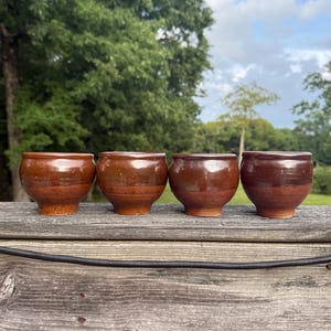 Könnte beinhalten: Vier kleine, abgerundete Keramikbecher in einer Reihe. Jeder Becher hat eine zweifarbige Glasur mit einem glänzenden, dunkelbraunen Finish im oberen Bereich und einem matten, rotbraunen, strukturierten Finish am unteren Sockel.