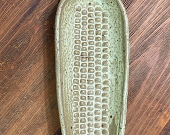 Può includere: Un piatto in ceramica verde chiaro a forma di pannocchia con finitura smaltata maculata e una trama a chicchi in rilievo. Il vassoio ovale allungato presenta un piccolo foro nella parte superiore con un cordino intrecciato color beige.