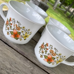 Könnte beinhalten: Vier weiße Keramikbecher mit Blumendesigns. Die Becher haben einen Henkel und zeigen ein Muster aus orangefarbenen, gelben und grünen Blumen.