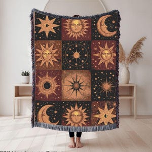 May include: A woven cotton throw blanket featuring a celestial design with suns, moons, and stars in shades of gold, brown, and black. The blanket has a fringed edge and the text "100% Handwoven Cotton" at the bottom.