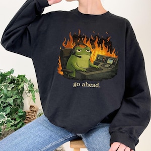 May include: A black crewneck sweatshirt featuring a graphic of a green frog wearing a headset, sitting at a desk with a computer, surrounded by orange and yellow flames. Below the graphic, the text "go ahead." is printed in a light color.