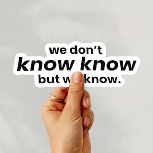 May include: A white sticker with black text that reads "we don't know know but w know." The sticker is held by a hand with a gold ring. The background is a plain, light gray color.