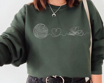 May include: A dark green sweatshirt with a white line drawing of a ball of yarn, a heart, and an open book. The sweatshirt is cropped and has a relaxed fit.