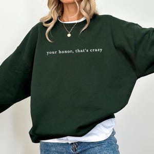 May include: A dark forest green crewneck sweatshirt featuring the white embroidered text "your honor, that's crazy" centred on the chest. The garment has long sleeves, a ribbed neckline, and a relaxed fit, worn over a white undershirt.