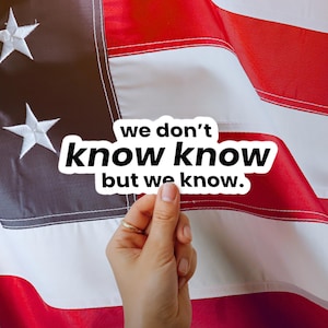 May include: A white sticker with the text "we don't know know but we know" in a black font is held in front of an American flag. The flag has red and white stripes with a dark blue field containing white stars.