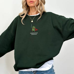 May include: Dark green sweatshirt with a white undershirt and blue jeans. The sweatshirt features a cartoon frog wearing a cape and holding a sword, with the text "DISSERTATION SURVIVOR" below the frog.