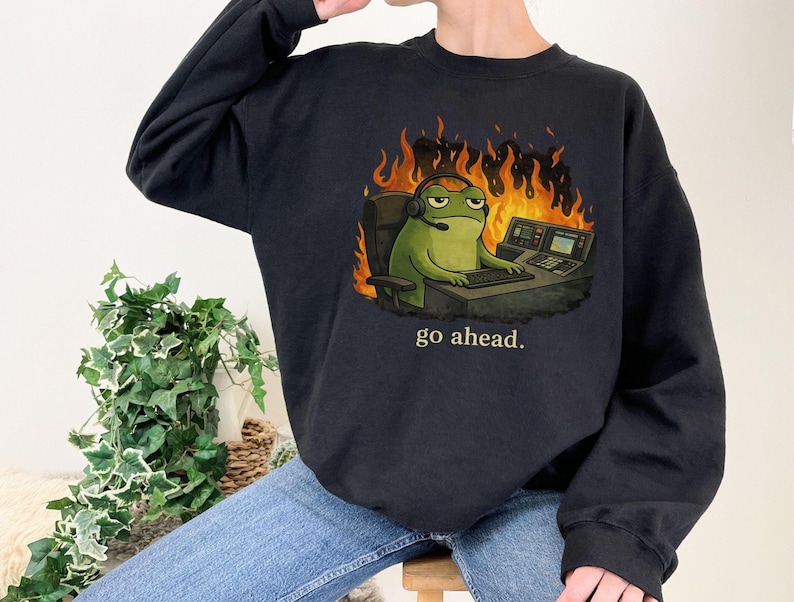 May include: A black crewneck sweatshirt featuring a graphic of a green frog wearing a headset, sitting at a desk with a computer, surrounded by orange and yellow flames. Below the graphic, the text "go ahead." is printed in a light color.