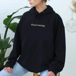 May include: Black hooded sweatshirt with the text "Good energy." in a cursive font. The sweatshirt has a front pocket and a drawstring hood. The model is wearing blue jeans.