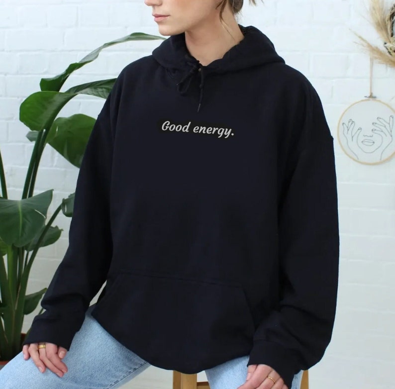 May include: Black hooded sweatshirt with the text "Good energy." in a cursive font. The sweatshirt has a front pocket and a drawstring hood. The model is wearing blue jeans.