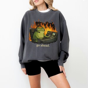 May include: A charcoal gray crewneck sweatshirt featuring a graphic of a green frog wearing a headset while sitting at a computer desk surrounded by orange flames. The text "go ahead." is printed in a light yellow font below the illustration.