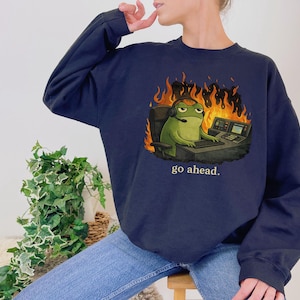 May include: A navy blue crewneck sweatshirt featuring a graphic of a green frog wearing a headset, sitting at a desk with a computer, surrounded by orange and yellow flames, with the text "go ahead." printed in white below the illustration.