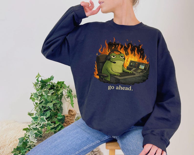 May include: A navy blue crewneck sweatshirt featuring a graphic of a green frog wearing a headset, sitting at a desk with a computer, surrounded by orange and yellow flames, with the text "go ahead." printed in white below the illustration.