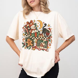 May include: An off-white short-sleeved t-shirt featuring a colourful Christmas-themed graphic. The design includes a snowman in a gift box, a black cat wearing a Santa hat, a gingerbread man, a snowy cabin scene, a crescent moon, and stars.