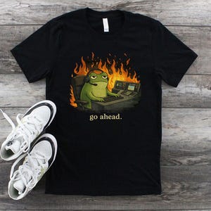 May include: Black t-shirt featuring a cartoon frog wearing a headset and sitting at a computer desk with flames in the background. The text "go ahead." is printed below the frog. White sneakers with black accents are next to the shirt.