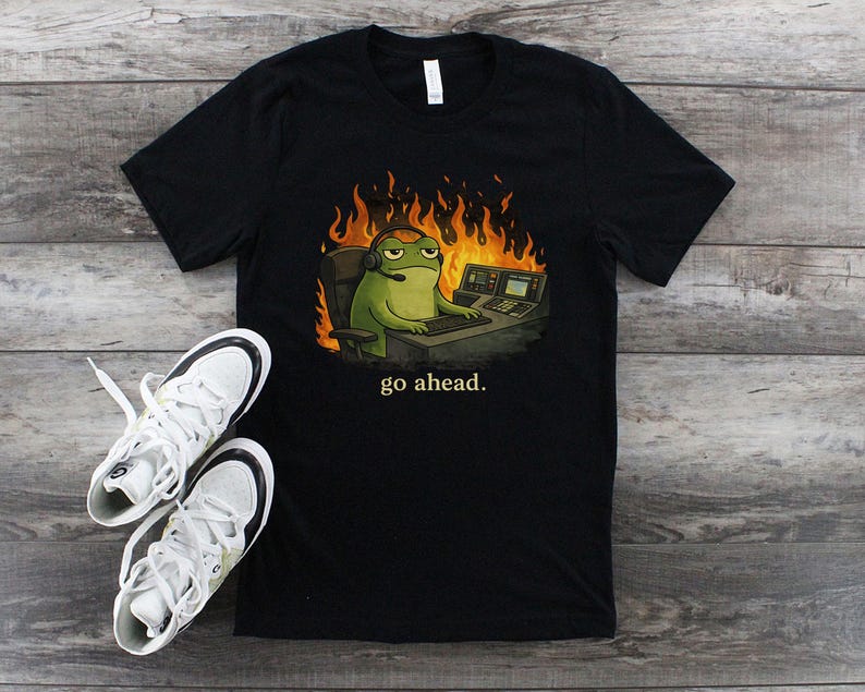 May include: Black t-shirt featuring a cartoon frog wearing a headset and sitting at a computer desk with flames in the background. The text "go ahead." is printed below the frog. White sneakers with black accents are next to the shirt.