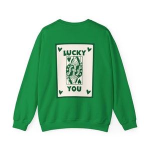 May include: A green sweatshirt featuring a playing card design on the back. The card depicts a queen with the words "LUCKY YOU" in green. The design is framed by green hearts.