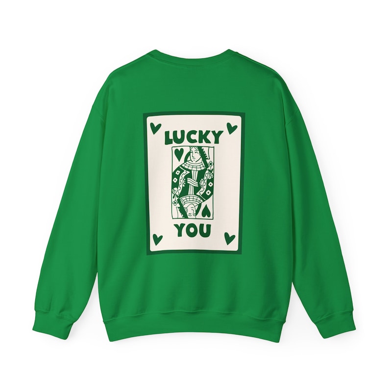 May include: A green sweatshirt featuring a playing card design on the back. The card depicts a queen with the words "LUCKY YOU" in green. The design is framed by green hearts.