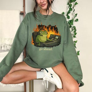 May include: An olive green crewneck sweatshirt featuring a graphic of a green frog wearing a headset, sitting at a desk with computer equipment in front of orange flames, with the text "go ahead." printed in small yellow letters below the graphic.