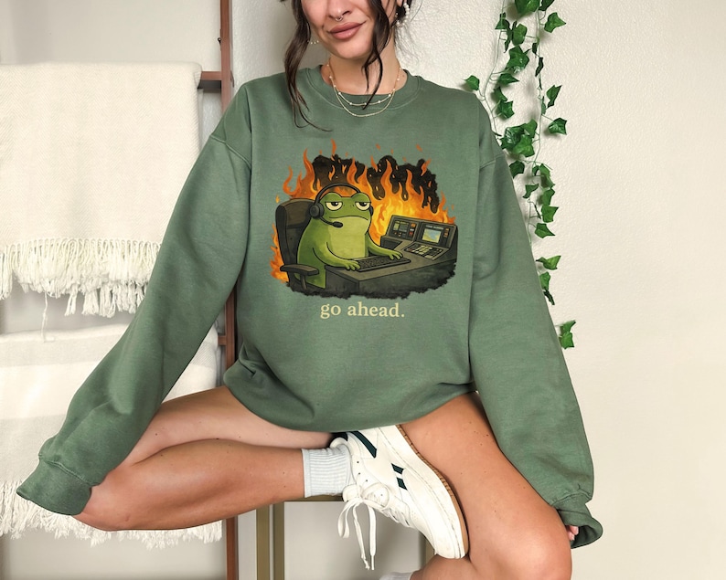 May include: An olive green crewneck sweatshirt featuring a graphic of a green frog wearing a headset, sitting at a desk with computer equipment in front of orange flames, with the text "go ahead." printed in small yellow letters below the graphic.