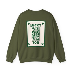May include: Olive green sweatshirt with a playing card design on the back. The card features a queen with the words "LUCKY YOU" in green. Green hearts are placed around the card. The sweatshirt is a solid color.