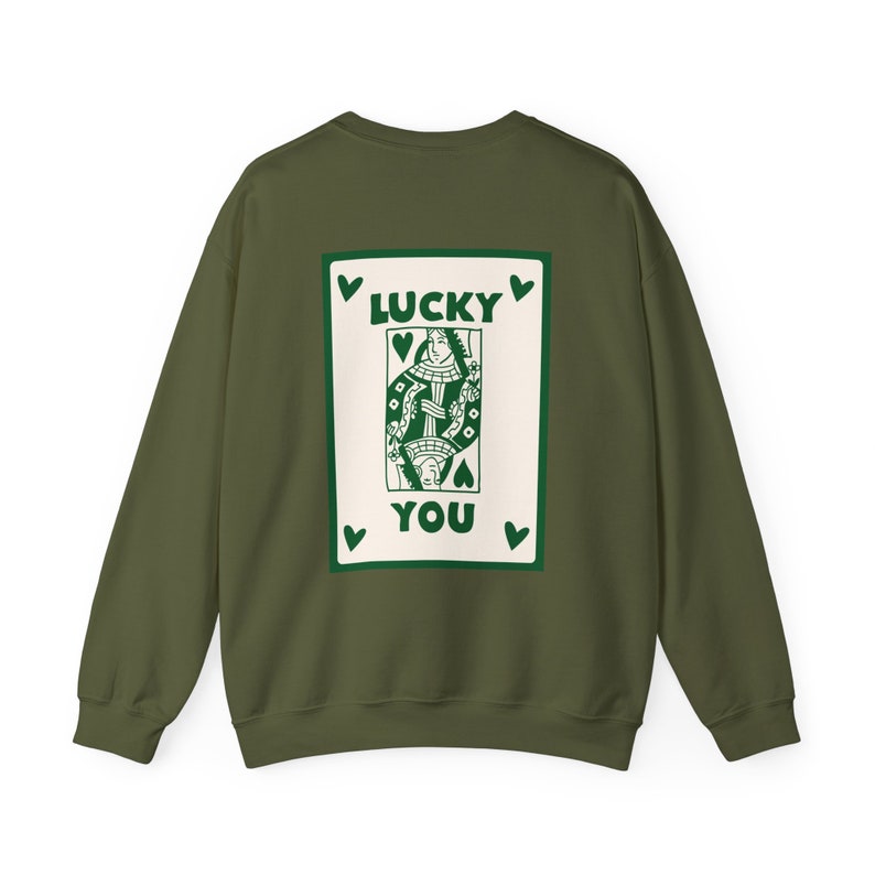 May include: Olive green sweatshirt with a playing card design on the back. The card features a queen with the words "LUCKY YOU" in green. Green hearts are placed around the card. The sweatshirt is a solid color.