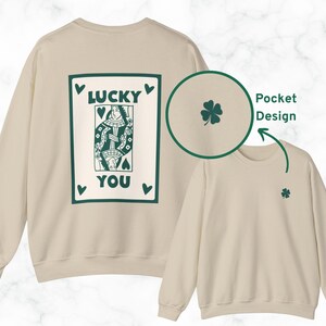 May include: A cream-colored sweatshirt with a green playing card design on the back that reads "LUCKY YOU." A small green four-leaf clover is on the front. The sweatshirt has a pocket design.