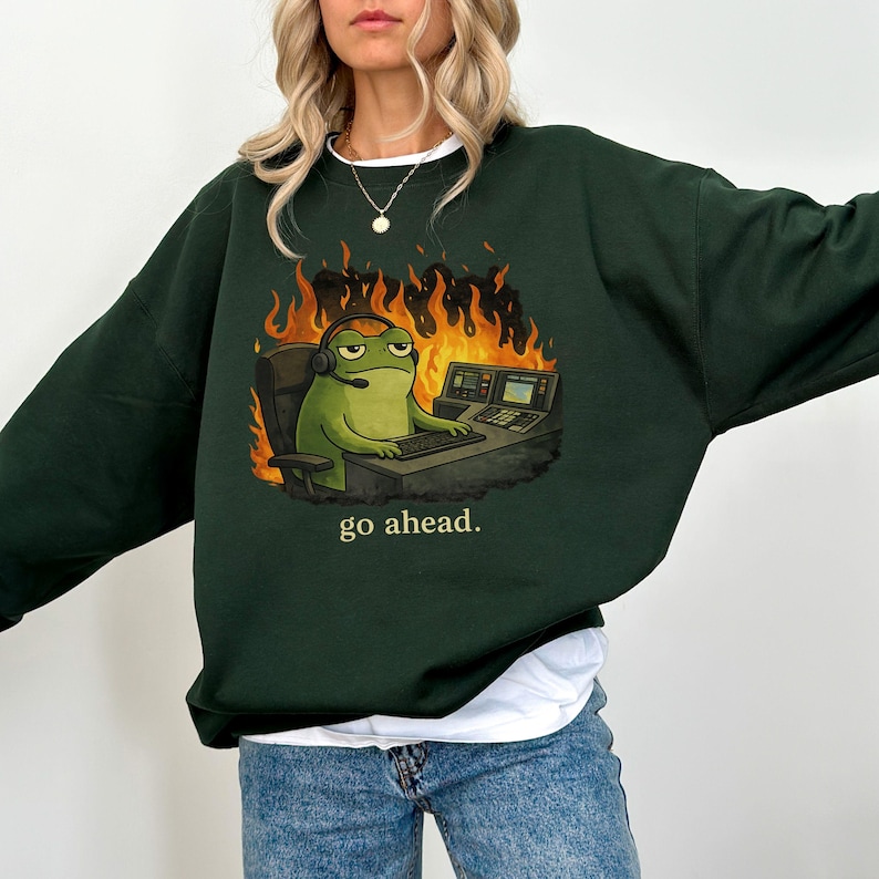 May include: A dark forest green crewneck sweatshirt featuring a graphic of a green frog wearing a headset, sitting at a computer desk in front of orange flames. The text "go ahead." is printed in a light yellow font below the graphic illustration.