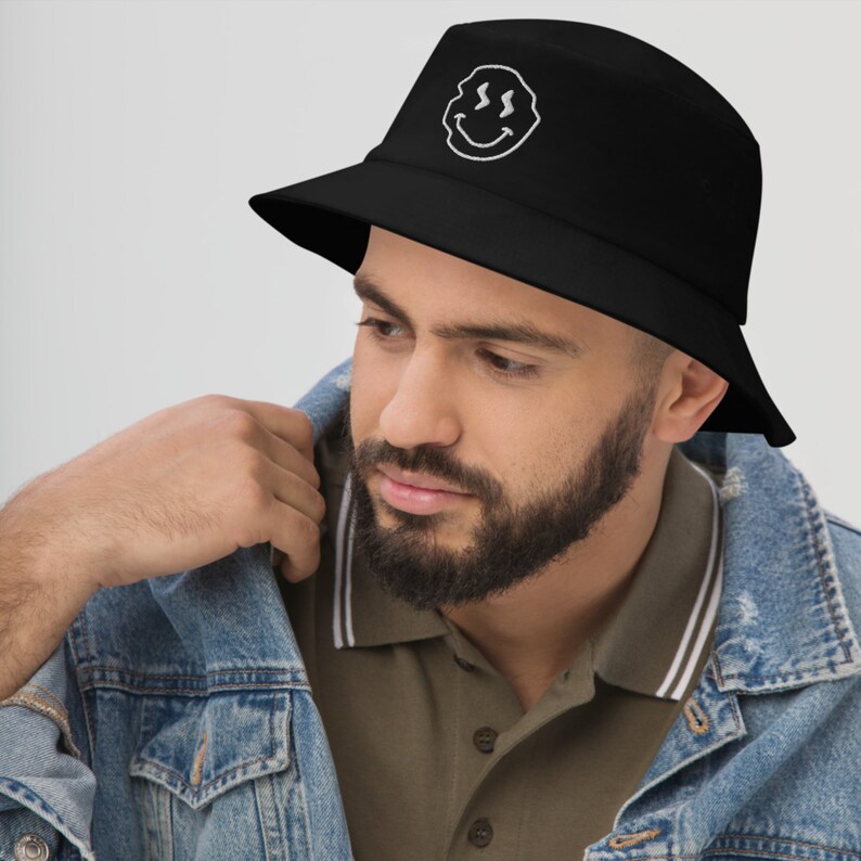 May include: A black bucket hat with a white embroidered smiley face. The hat has a curved brim and a relaxed fit.