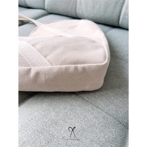 K&ouml;nnte beinhalten: Hellbeige Canvas-Umh&auml;ngetasche mit rechteckiger Basis und abgerundeter Oberseite. Die Tasche hat einen passenden Riemen und liegt auf einem hellblau-gr&uuml;nen Sofa. Die Textur der Tasche ist sichtbar, und die Gesamt&auml;sthetik ist minimalistisch.