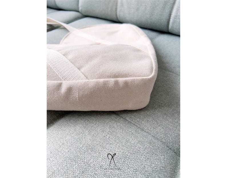 K&ouml;nnte beinhalten: Hellbeige Canvas-Umh&auml;ngetasche mit rechteckiger Basis und abgerundeter Oberseite. Die Tasche hat einen passenden Riemen und liegt auf einem hellblau-gr&uuml;nen Sofa. Die Textur der Tasche ist sichtbar, und die Gesamt&auml;sthetik ist minimalistisch.