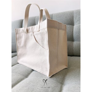 May include: A large, off-white canvas tote bag with a single, large pocket on the front. The bag has two fabric handles.