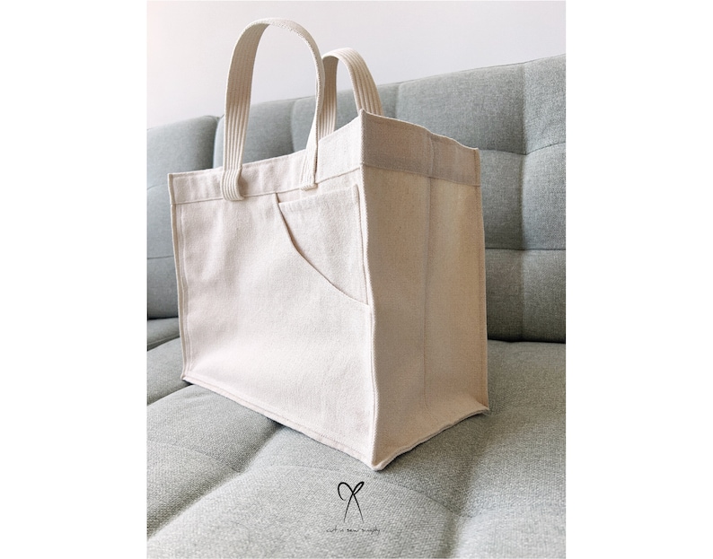 May include: A large, off-white canvas tote bag with a single, large pocket on the front. The bag has two fabric handles.